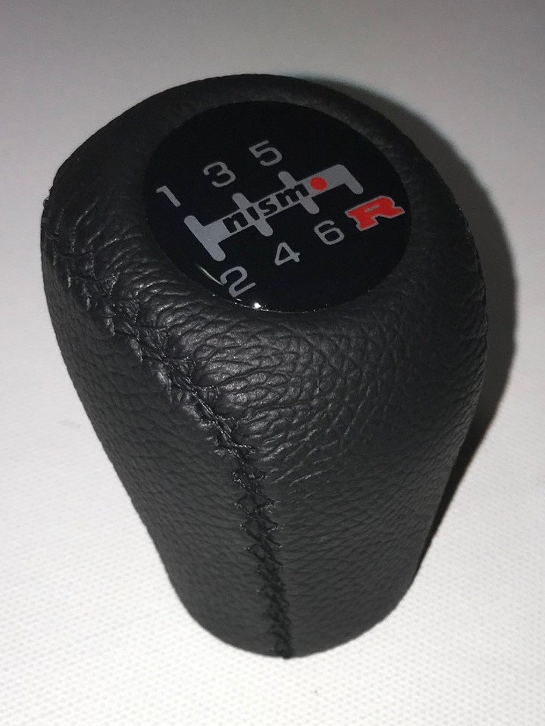 May include: Black leather gear shift knob with a black and silver Nismo logo and red R on a black background. The knob has a stitched seam and numbers 1 through 6.