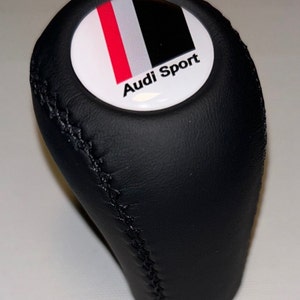 May include: Black leather gear shift knob with a white, red, and black emblem that reads "Audi Sport". The knob has a stitched seam and a rounded top.