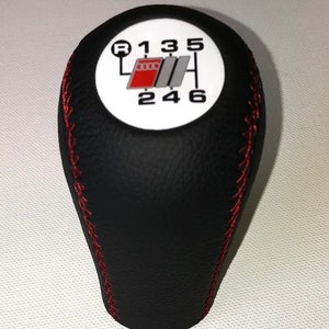 Könnte beinhalten: Schwarzer Leder-Schalthebelknauf mit roten Nähten und einer weißen runden Oberseite mit den Zahlen 1, 3, 5, R, 2, 4 und 6 darauf gedruckt. Die Oberseite hat auch ein kleines Logo mit den Buchstaben "Audi" darauf gedruckt.