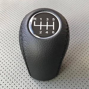Könnte beinhalten: Schwarzer Leder-Schalthebelknauf mit einem silbernen Ring und weißen Zahlen 1, 2, 3, 4, 5, 6 und R.