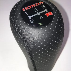 Op de afbeelding: Zwarte leren Honda versnellingspookknop met witte stiksels en een geperforeerd ontwerp. De bovenkant heeft een zwarte wijzerplaat met het Honda-logo in rood en witte cijfers en versnellingsindicatoren.