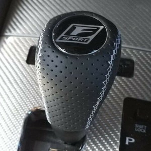 May include: A black leather gear shift knob with a silver F SPORT logo. The knob has a perforated design and white stitching.