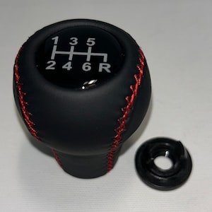 May include: A black gear shift knob with red stitching and a glossy top insert displaying a white 6-speed manual transmission shift pattern with numbers 1 through 6 and R for reverse. A separate black circular plastic mounting ring is included.