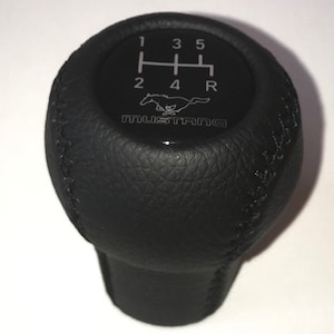 May include: Black leather gear shift knob with white stitching and a silver Mustang logo with the numbers 1, 2, 3, 4, 5, and R.