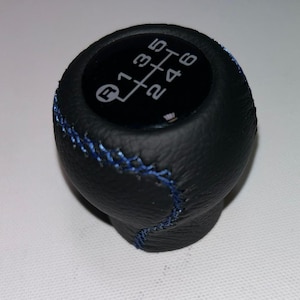 Op de afbeelding: Zwarte versnellingspookknop met blauwe stiksels. De bovenkant van de knop heeft een diagram van het schakelpatroon, met witte cijfers en letters. De knop is ontworpen voor een handgeschakelde versnellingsbak.