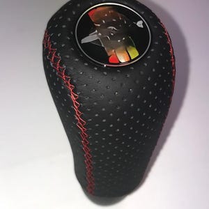 May include: Black perforated leather gear shift knob with red stitching. The top has a circular emblem with a colourful design. The knob is designed for a vehicle's manual transmission and is set against a white background.