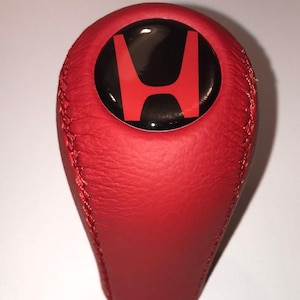 Könnte beinhalten: Roter Lederschaltknauf mit einem schwarzen und roten Honda-Logo. Der Schaltknauf hat eine leuchtend rote Farbe und eine genähte Naht. Das Honda-Logo befindet sich mittig auf der Oberseite des Knaufs.