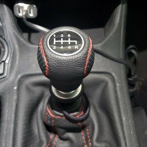 May include: A black gear stick knob with red stitching. The shift pattern is visible on the top, with numbers and the letter "R" for reverse. The knob is surrounded by a black leather boot with red stitching.