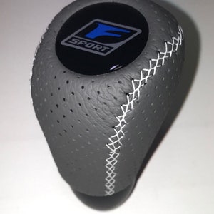 Peut inclure: Pommeau de levier de vitesses en cuir gris avec surpiqûres blanches. Le dessus présente un emblème noir avec le mot "SPORT" et un logo "F" bleu. Le pommeau a une texture perforée pour l'adhérence.
