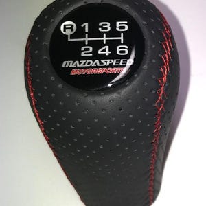 May include: Black leather gear stick knob with red stitching. The top displays a gear shift pattern with the numbers 1, 2, 3, 4, 5, 6, and R. The text "MAZDASPEED MOTORSPORTS" is also visible.