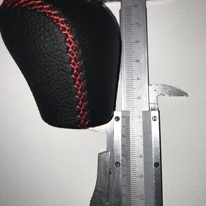 May include: A black leather gear shift knob with red stitching, measured with a caliper to be approximately 6.5 inches in length.