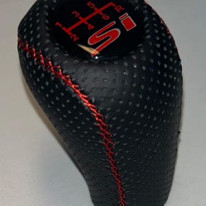 Peut inclure: Pommeau de levier de vitesses noir avec des coutures rouges et un indicateur de schéma de vitesses. Le dessus du pommeau comporte un emblème rouge et noir avec la lettre "S" et les numéros de vitesse.