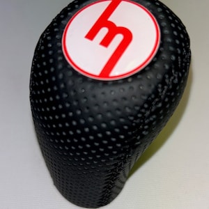 Könnte beinhalten: Schwarzer Leder-Schalthebelknauf mit einem roten "M"-Logo in einem weißen Kreis.