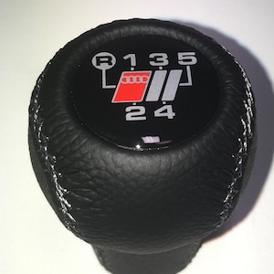 May include: Black leather gear shift knob with white stitching and a black and silver gear shift pattern with the Audi logo in red. The gear shift pattern shows the numbers 1, 3, 5, R, 2, and 4.