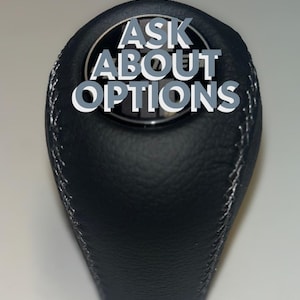May include: Black leather gear shift knob with white stitching. The text "ASK ABOUT OPTIONS" is displayed in white and gray lettering on the top of the knob. The knob has a textured surface.