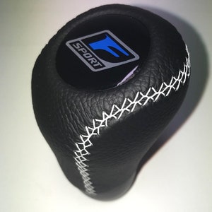 Könnte beinhalten: Schwarzer Lederschaltknauf mit weißen Nähten. Die Oberseite hat ein schwarzes Emblem mit dem Wort "SPORT" und einem blauen "F"-Logo. Der Knauf hat eine strukturierte Oberfläche und eine abgerundete Form.