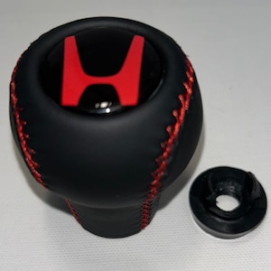 May include: A black spherical gear shift knob featuring a red Honda logo on the top centre. The knob is wrapped in a matte black material with prominent red cross-stitching running vertically along the sides. A separate black plastic ring is included.