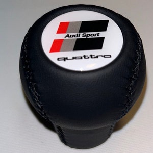 Könnte beinhalten: Schwarzer Leder-Schalthebelknauf mit einem weißen runden Emblem mit dem Audi Sport Logo und dem Wort "quattro" in weißen Buchstaben.