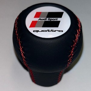 Könnte beinhalten: Schwarzer Leder-Schalthebelknauf mit roten Nähten und einem weißen runden Emblem, auf dem "Audi Sport" und "quattro" in schwarzen Buchstaben stehen.