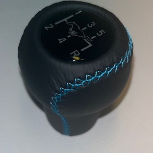 May include: Black leather gear shift knob with blue stitching. The top features a black face with white numbers and a running horse logo. The gear pattern is visible. A stylish upgrade for a vehicle's interior.