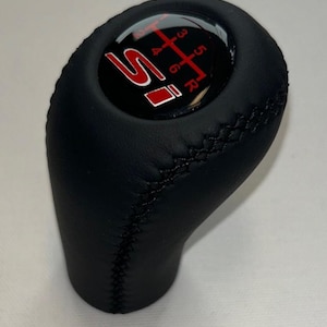 Op de afbeelding: Zwarte leren versnellingspookknop met rood "Si" logo en schakelpatroon. Het schakelpatroon omvat de versnellingen 1, 2, 3, 4, 5, 6 en R (achteruit). De knop heeft een gestikte naad en een glanzende bovenkant.
