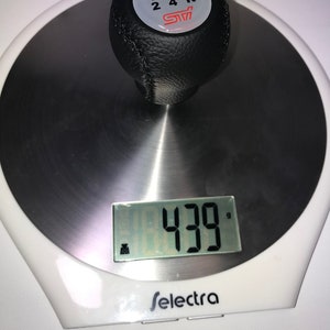 May include: A black leather gear shift knob with a white gear shift pattern and the text "STI" in red. The knob is on a digital scale that reads 439 grams.