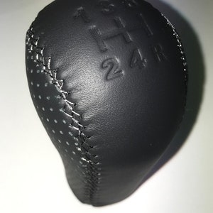 May include: Black leather gear shift knob with white stitching and a pattern of small holes. The knob has the numbers 1, 3, 5, and 6, and the letter H, along with the numbers 2, 4, and R embossed on the top.