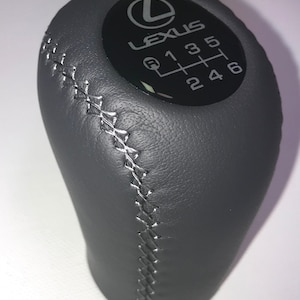May include: A black leather gear shift knob with a silver stitching detail. The knob has a black circular emblem with the word "Lexus" and a 6-speed gear pattern.