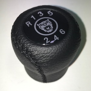 Può includere: Pomello del cambio in pelle nera con un emblema di giaguaro argentato e numeri bianchi 1, 3, 5, R, 2, 4, 6.