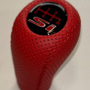 Puede incluir: Un pomo de cambio de cuero rojo perforado con una parte superior negra. La parte superior presenta las letras "SI" en blanco y rojo, junto con un patrón de cambio de marchas. El pomo está diseñado para una transmisión manual.
