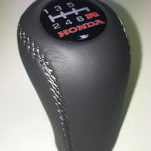 Op de afbeelding: Een zwarte versnellingspookknop met witte stiksels. De bovenkant van de knop heeft een zwarte wijzerplaat met het versnellingspatroon en het woord "HONDA" in het rood. Het versnellingspatroon bevat de letter "R" voor achteruit.