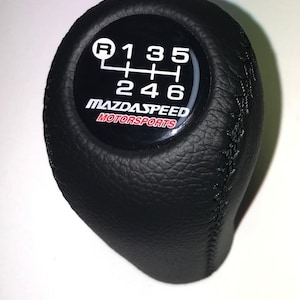 Könnte beinhalten: Schwarzer Lederschaltknauf mit weißem Gangmuster und dem Text "MAZDA SPEED MOTORSPORTS" in Rot und Weiß. Das Gangmuster umfasst R, 1, 2, 3, 4, 5 und 6.