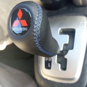 Może przedstawiać: Czarny skórzany gałka zmiany biegów z niebieskimi szwami i logo Mitsubishi. Gałka jest przymocowana do srebrnej dźwigni zmiany biegów.