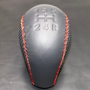 Può includere: Un pomello del cambio nero con cuciture rosse. La parte superiore del pomello presenta in rilievo lo schema delle marce: 1, 3, 5, H, 2, 4 e R. Il pomello ha una trama perforata sui lati.