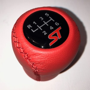Op de afbeelding: Rode leren versnellingspookknop met zwart en zilver "ST" logo en een zwart versnellingschema met witte nummers 1, 2, 3, 4, 5, 6 en R.