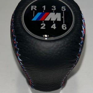 Peut inclure: Un pommeau de levier de vitesse en cuir noir avec un schéma de transmission manuelle à six vitesses incluant la marche arrière. Le centre affiche le logo M en bleu, violet et rouge. Les côtés présentent des coutures décoratives tricolores.