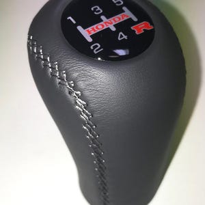 May include: Black gear stick knob with silver stitching. The top has a black circle with white numbers and the word "HONDA" in red. The gear pattern is visible, with "R" in red. A car accessory.