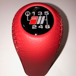 May include: Red leather gear shift knob with a black and silver gear shift pattern and the Audi logo. The gear shift pattern shows the numbers 1, 3, 5, 2, 4, 6, and R.