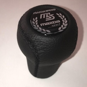 May include: Black leather gear stick knob featuring the text "MAZDASPEED" and "MS MAZDA sport" in white. The knob has a stitched seam and a textured surface.