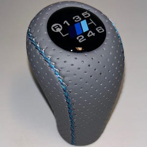 May include: Grey perforated leather gear knob with blue stitching. The top has a black circle displaying the gear shift pattern in white and blue. The pattern includes R, 1, 2, 3, 4, 5, and 6.