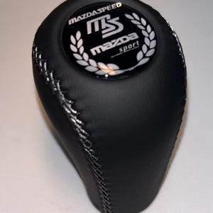 May include: Black leather gear stick knob with silver stitching. The top has a glossy emblem with the text "MAZDASPEED", "MS", and "MAZDA sport" in white, surrounded by a laurel wreath. Designed for a vehicle.