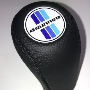 May include: Black leather gear shift knob with a white circle and blue stripes, with the text "4RUNNER" in black lettering.
