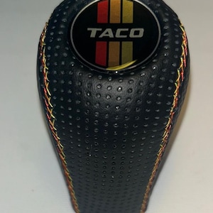 Può includere: Pomello del cambio nero con la scritta "TACO" in bianco su un emblema circolare con strisce rosse, gialle e verdi. Il pomello ha una superficie strutturata ed è cucito con filo giallo e rosso.