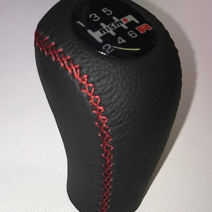 Puede incluir: Pomo de cambio de marchas de cuero negro con costuras rojas y un logotipo Nismo en blanco y negro con una "R" roja en la parte superior.