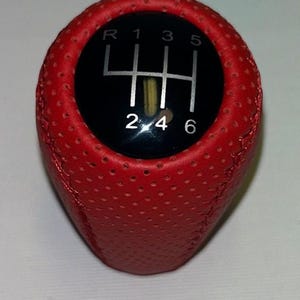 Può includere: Pomello del cambio in pelle rossa traforata. La parte superiore presenta un cerchio nero con le marce: R, 1, 3, 5, 2, 4 e 6. Progettato per un cambio manuale.