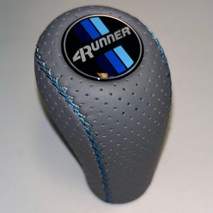 May include: Gray perforated shift knob with blue stitching. The knob features a circular emblem with the text "4Runner" and blue stripes. The shift knob is designed for a vehicle.