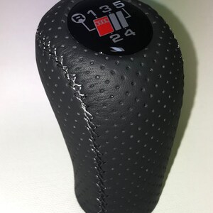 May include: Black leather gear shift knob with a black and red gear shift pattern. The pattern includes the numbers 1, 2, 3, 4, 5, and R. The knob has a perforated surface and white stitching.