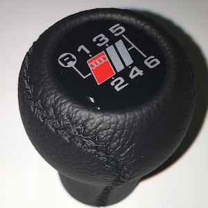 Può includere: Pomello del cambio in pelle nera con uno schema di cambio nero e argento e un logo Audi rosso. I numeri 1, 3, 5, R, 2, 4 e 6 sono stampati sul pomello.