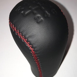 Può includere: Pomello del cambio in pelle nera con cuciture rosse. Il motivo delle marce è impresso sulla parte superiore. Le cuciture rosse corrono verticalmente lungo il lato del pomello.