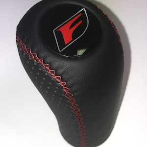 Könnte beinhalten: Schwarzer Leder-Schalthebelknauf mit roten Nähten und einem roten "F"-Logo.